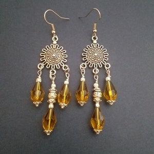 May include: A pair of gold tone dangle earrings with amber glass teardrop beads. The earrings feature a decorative metal charm with a circular design.
