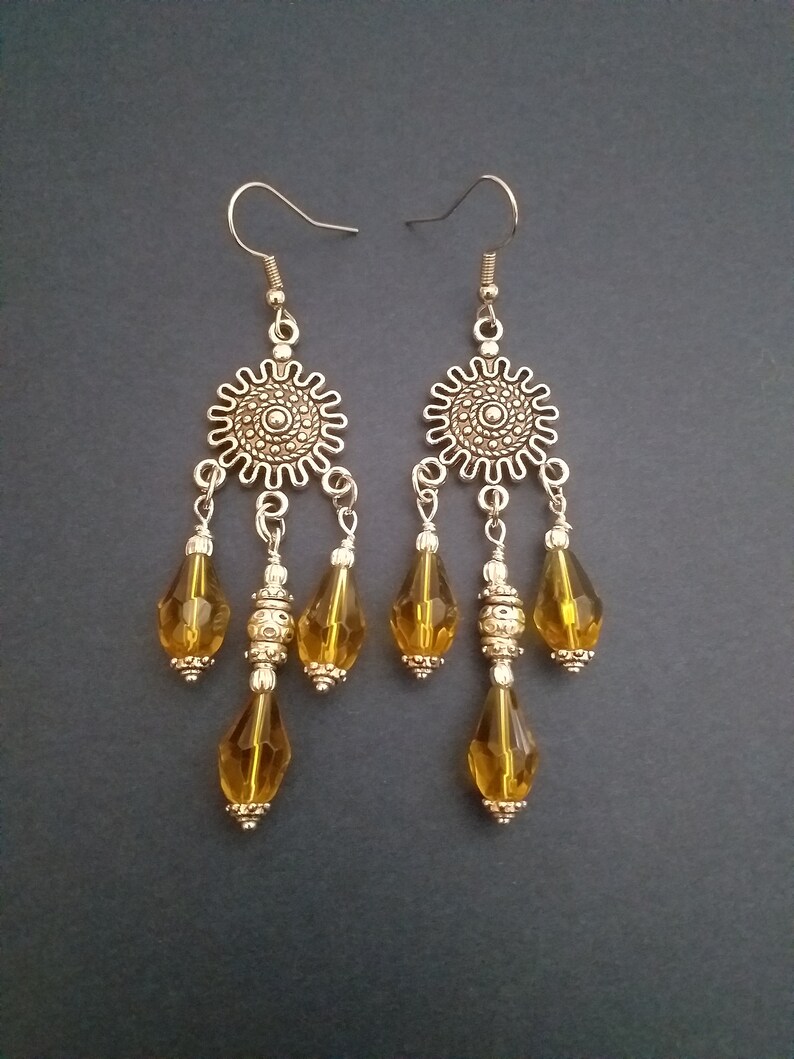 May include: A pair of gold tone dangle earrings with amber glass teardrop beads. The earrings feature a decorative metal charm with a circular design.