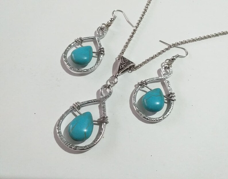 K&ouml;nnte beinhalten: Ein silbernes Halsketten- und Ohrring-Set mit t&uuml;rkisfarbenen Tr&auml;nensteinen. Die Halskette hat einen Anh&auml;nger mit einem keltischen Knoten und die Ohrringe sind im H&auml;nge-Stil.