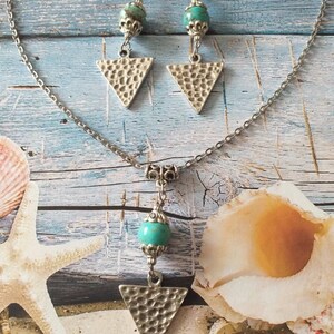 May include: A silver chain necklace with a turquoise bead and a hammered silver triangle pendant. Matching earrings with turquoise beads and hammered silver triangle pendants.