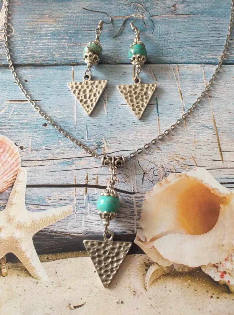 May include: A silver chain necklace with a turquoise bead and a hammered silver triangle pendant. Matching earrings with turquoise beads and hammered silver triangle pendants.