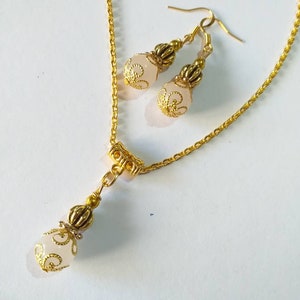 May include: A gold chain necklace with a pendant and matching earrings. The pendant and earrings feature a white, round bead with a gold filigree design.