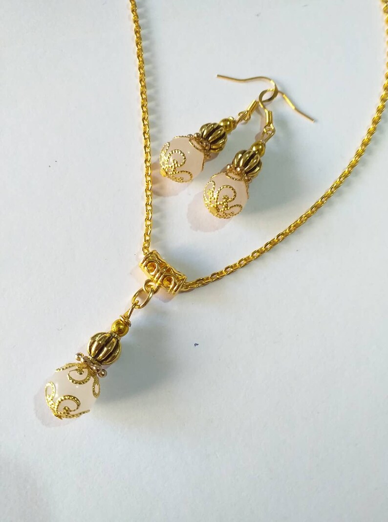 May include: A gold chain necklace with a pendant and matching earrings. The pendant and earrings feature a white, round bead with a gold filigree design.