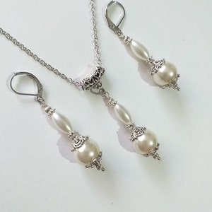May include: A silver necklace and earrings set with white pearl accents. The necklace features a silver chain with a decorative pendant and two white pearls. The earrings have a silver hook and two white pearls.