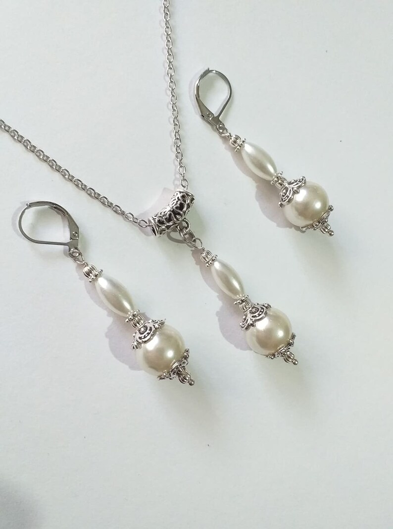 May include: A silver necklace and earrings set with white pearl accents. The necklace features a silver chain with a decorative pendant and two white pearls. The earrings have a silver hook and two white pearls.