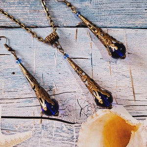 May include: A set of gold-tone jewelry with blue glass teardrop pendants. The necklace has a chain link design and the earrings have a hook closure.