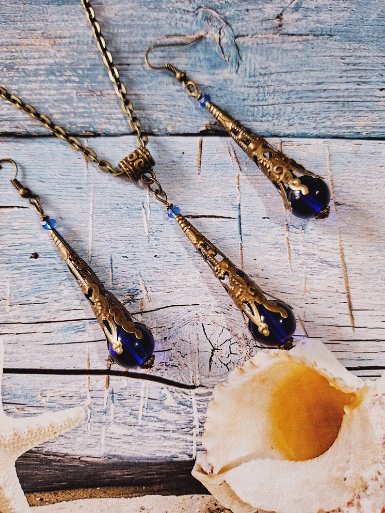 May include: A set of gold-tone jewelry with blue glass teardrop pendants. The necklace has a chain link design and the earrings have a hook closure.