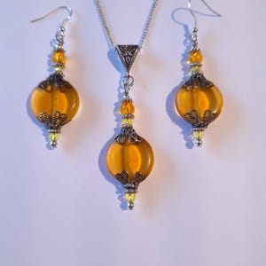 May include: A set of amber glass and silver tone earrings and necklace. The earrings and necklace feature a round amber glass bead with a silver tone filigree design.