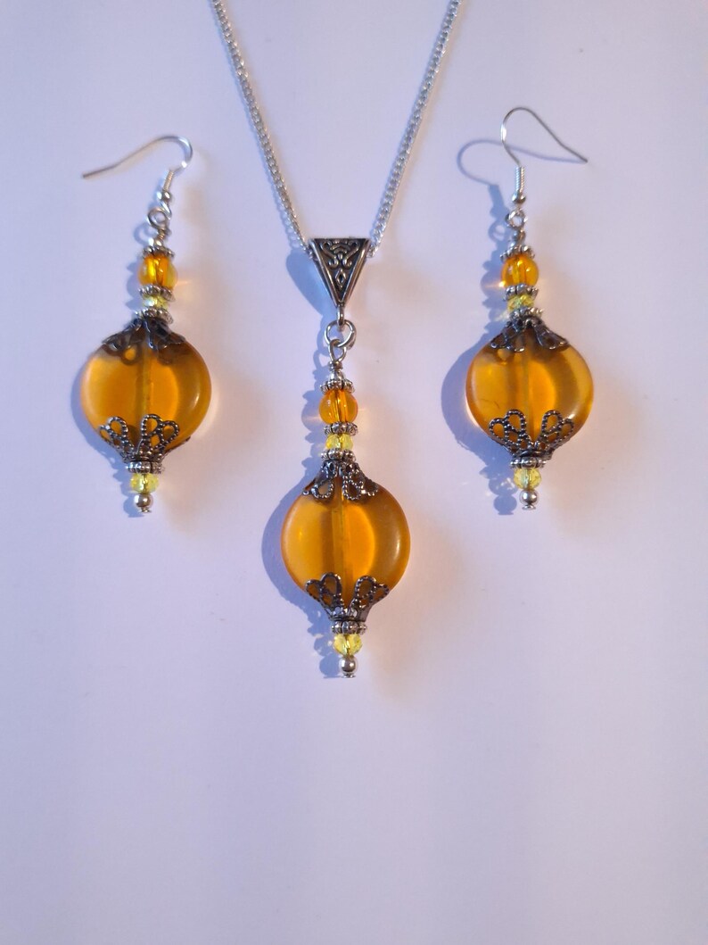 May include: A set of amber glass and silver tone earrings and necklace. The earrings and necklace feature a round amber glass bead with a silver tone filigree design.