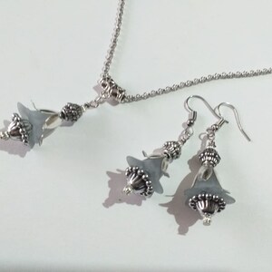 May include: A silver necklace and earrings set with grey flower-shaped pendants. The necklace has a delicate chain and the earrings have silver hooks.