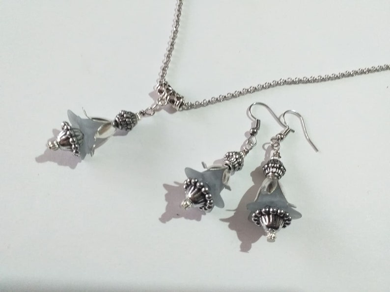 May include: A silver necklace and earrings set with grey flower-shaped pendants. The necklace has a delicate chain and the earrings have silver hooks.