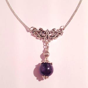 Puede incluir: Un collar de cadena de plata con un colgante de plata con un dise&ntilde;o floral. El colgante est&aacute; unido a una piedra preciosa de color p&uacute;rpura oscuro.