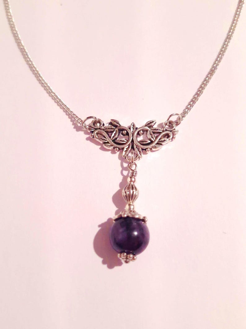 Puede incluir: Un collar de cadena de plata con un colgante de plata con un dise&ntilde;o floral. El colgante est&aacute; unido a una piedra preciosa de color p&uacute;rpura oscuro.