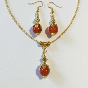 K&ouml;nnte beinhalten: Eine goldene Kettenkette mit einem roten Edelstein-Anh&auml;nger und passenden Ohrringen. Anh&auml;nger und Ohrringe sind mit verzierten goldenen Akzenten versehen.