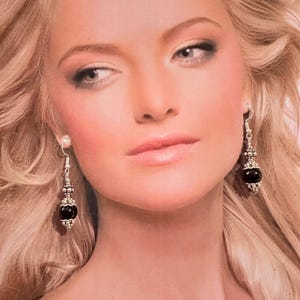 May include: Silver dangle earrings with a black, round bead at the bottom. The earrings have a decorative silver design with a small, clear stone at the top. The earrings are shown on a person with blonde hair.