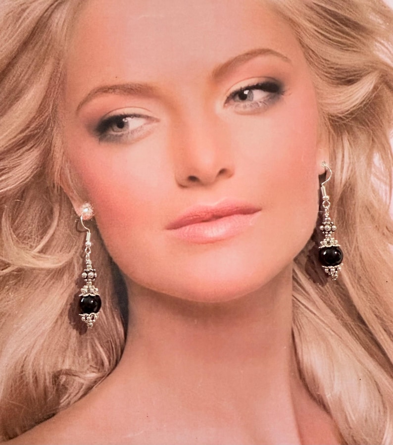 May include: Silver dangle earrings with a black, round bead at the bottom. The earrings have a decorative silver design with a small, clear stone at the top. The earrings are shown on a person with blonde hair.