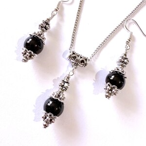 May include: A jewelry set featuring a necklace and matching earrings. The set includes a silver-toned chain and earring hooks. Each piece has a large, round, black bead accented with ornate silver-colored metalwork. The jewelry is displayed on a white background.