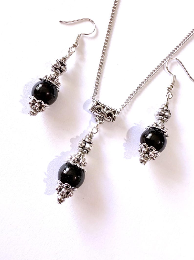May include: A jewelry set featuring a necklace and matching earrings. The set includes a silver-toned chain and earring hooks. Each piece has a large, round, black bead accented with ornate silver-colored metalwork. The jewelry is displayed on a white background.