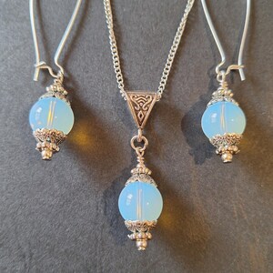 May include: A silver necklace and earrings set featuring light blue glass beads. The beads are round and have a silver filigree design around them. The necklace has a silver chain and a silver pendant with a Celtic knot design.