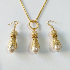 May include: A gold necklace with a circular pendant and matching earrings. The pendant and earrings feature a delicate filigree design and a white pearl drop.