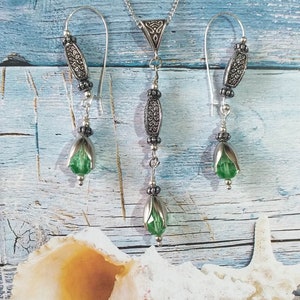 May include: A silver necklace and earrings set with green glass beads and silver filigree accents. The necklace has a pendant with a teardrop-shaped green glass bead.