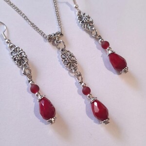 Puede incluir: Un collar y pendientes de plata con una cuenta roja en forma de l&aacute;grima y un colgante con dise&ntilde;o floral.