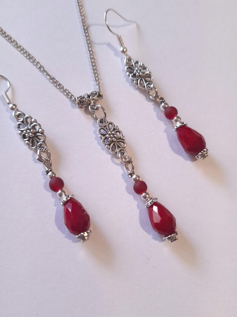 Puede incluir: Un collar y pendientes de plata con una cuenta roja en forma de l&aacute;grima y un colgante con dise&ntilde;o floral.