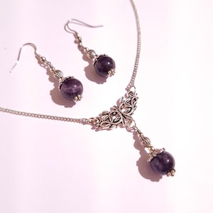 May include: A silver necklace with a delicate floral design and a dangling amethyst gemstone. Matching amethyst earrings with silver hooks are included.