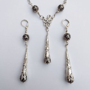 May include: A silver necklace with a dangling teardrop-shaped pendant and two matching earrings. The necklace and earrings feature black beads and intricate silver designs.