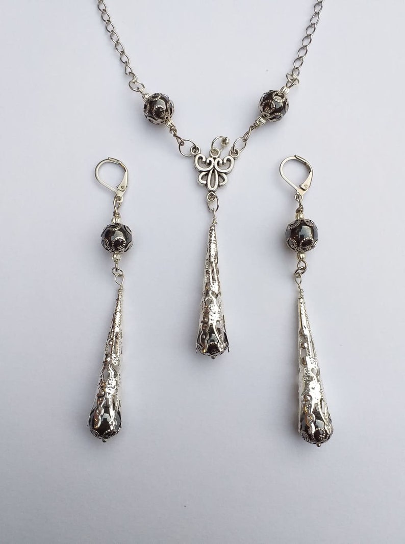 May include: A silver necklace with a dangling teardrop-shaped pendant and two matching earrings. The necklace and earrings feature black beads and intricate silver designs.