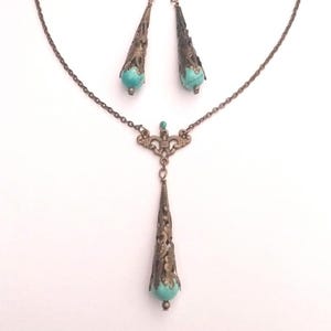 Puede incluir: Un conjunto de collar y pendientes de turquesa y bronce. El collar presenta una cadena delicada con un colgante que tiene una piedra turquesa y un dise&ntilde;o de filigrana intrincado. Los pendientes son pendientes colgantes con piedras turquesa y dise&ntilde;os de filigrana.