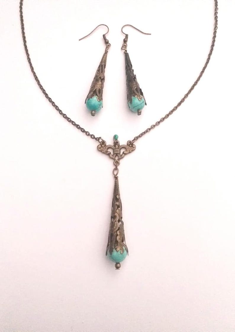 Puede incluir: Un conjunto de collar y pendientes de turquesa y bronce. El collar presenta una cadena delicada con un colgante que tiene una piedra turquesa y un dise&ntilde;o de filigrana intrincado. Los pendientes son pendientes colgantes con piedras turquesa y dise&ntilde;os de filigrana.