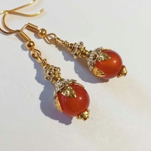 K&ouml;nnte beinhalten: Ein Paar goldene Ohrh&auml;nger mit orangefarbenen Karneolperlen und verzierten goldenen Akzenten.
