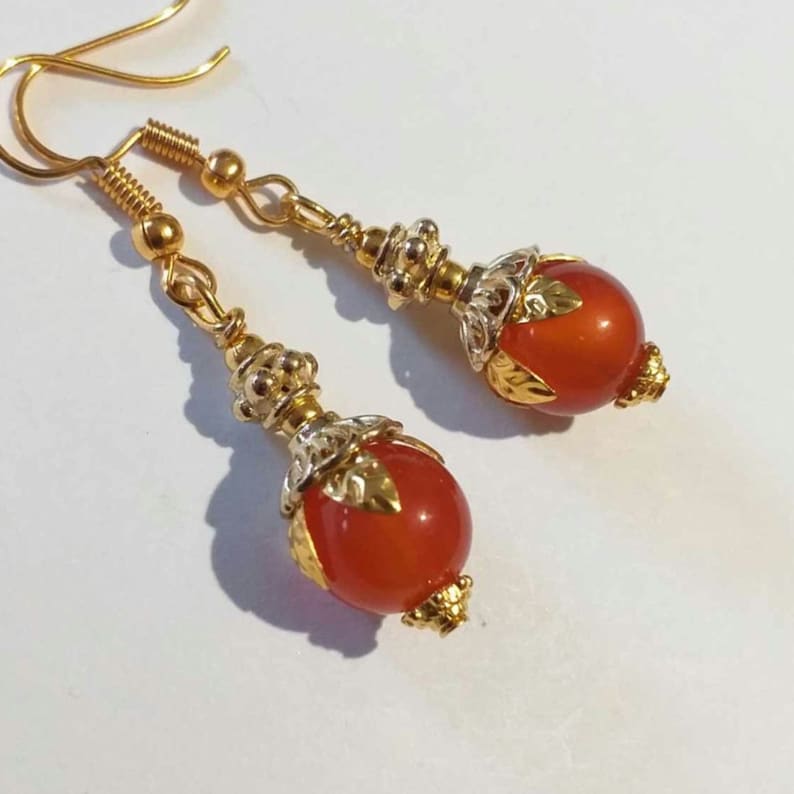 K&ouml;nnte beinhalten: Ein Paar goldene Ohrh&auml;nger mit orangefarbenen Karneolperlen und verzierten goldenen Akzenten.
