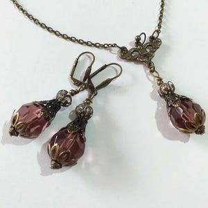 Op de afbeelding: Een set sieraden met een ketting met hanger en oorbellen. De ketting heeft een delicate ketting met een bronzen vlinderhanger en een enkele traanvormige paarse kristal. De oorbellen zijn ook van brons met traanvormige paarse kristallen.