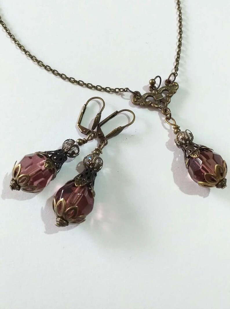 Op de afbeelding: Een set sieraden met een ketting met hanger en oorbellen. De ketting heeft een delicate ketting met een bronzen vlinderhanger en een enkele traanvormige paarse kristal. De oorbellen zijn ook van brons met traanvormige paarse kristallen.