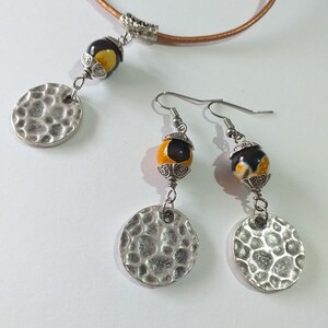May include: A jewelry set featuring a necklace and earrings. The necklace has a brown leather cord with a pendant of a black and yellow bead and a hammered silver disc. The earrings have matching beads and discs.