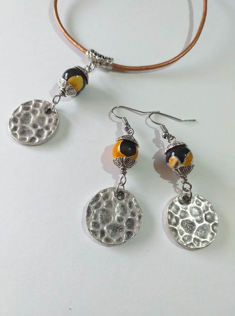 May include: A jewelry set featuring a necklace and earrings. The necklace has a brown leather cord with a pendant of a black and yellow bead and a hammered silver disc. The earrings have matching beads and discs.