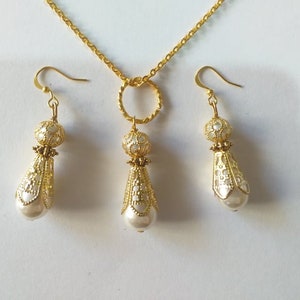 May include: A gold necklace with a teardrop-shaped pendant and matching earrings. The pendant and earrings are decorated with a white filigree design and feature a pearl at the bottom.