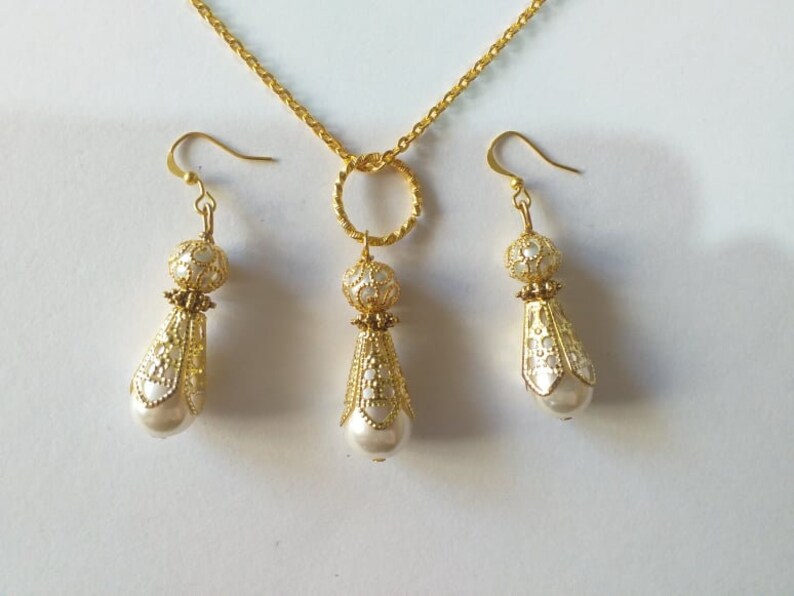 May include: A gold necklace with a teardrop-shaped pendant and matching earrings. The pendant and earrings are decorated with a white filigree design and feature a pearl at the bottom.