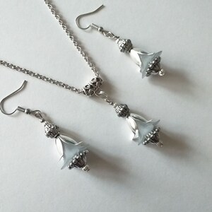 May include: A silver necklace and earrings set with a delicate chain and a white flower-shaped pendant. The pendant and earrings have a silver filigree design.