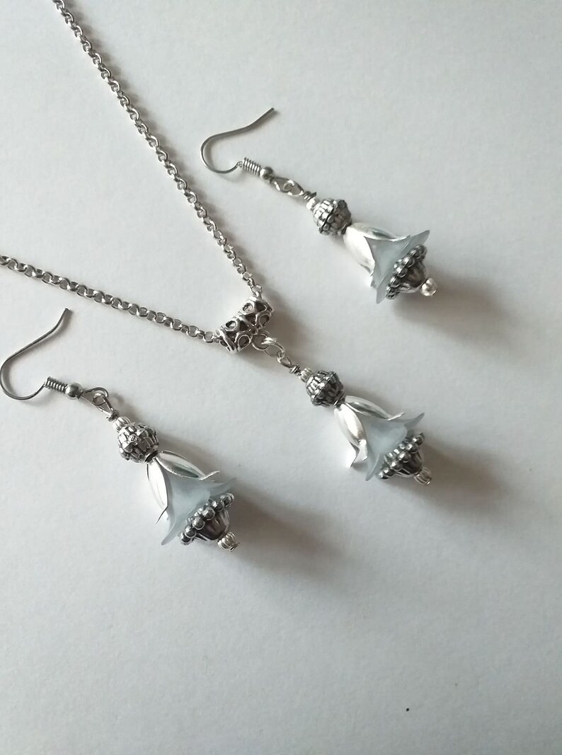 May include: A silver necklace and earrings set with a delicate chain and a white flower-shaped pendant. The pendant and earrings have a silver filigree design.