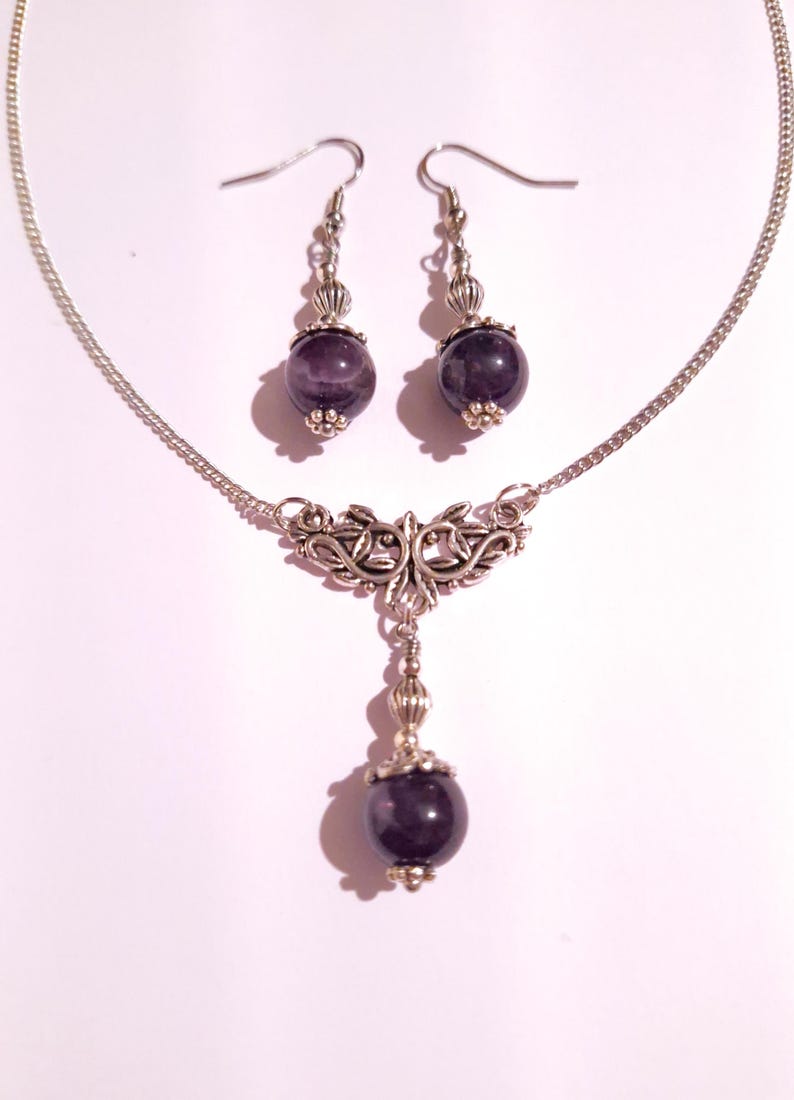 Puede incluir: Un conjunto de collar y pendientes de plata con un colgante de amatista p&uacute;rpura y pendientes a juego. El collar presenta un dise&ntilde;o de filigrana de plata.