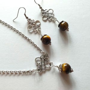 May include: A silver necklace and earrings set with a brown tiger's eye stone pendant and matching earrings. The pendant and earrings feature a silver, intricate, knot-like design.