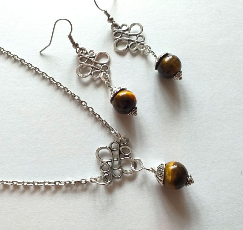 May include: A silver necklace and earrings set with a brown tiger's eye stone pendant and matching earrings. The pendant and earrings feature a silver, intricate, knot-like design.