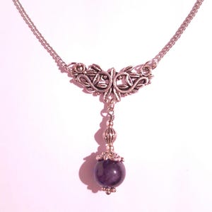 Puede incluir: Un collar de plata con un delicado dise&ntilde;o floral y una piedra preciosa de amatista colgante. El collar est&aacute; sobre un fondo blanco.