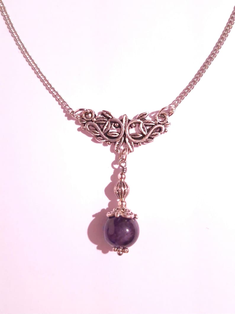 Puede incluir: Un collar de plata con un delicado dise&ntilde;o floral y una piedra preciosa de amatista colgante. El collar est&aacute; sobre un fondo blanco.