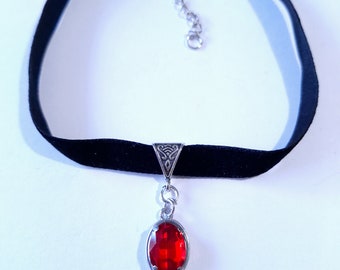 Puede incluir: Collar gargantilla de terciopelo negro con cadena y cierre plateados. El colgante presenta una gema roja de forma ovalada engastada en un marco de metal plateado. Fondo blanco.