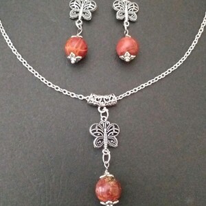 May include: A silver chain necklace with a red and brown stone pendant and a silver butterfly charm. Matching earrings with red and brown stone and silver butterfly charms.