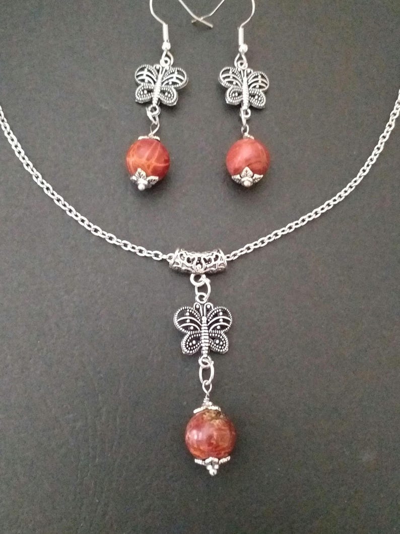 May include: A silver chain necklace with a red and brown stone pendant and a silver butterfly charm. Matching earrings with red and brown stone and silver butterfly charms.